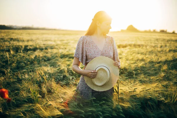 夕日の光の中で大麦のフィールドに立って花のドレスの美しい女性 スタイリッシュな女性は 麦藁帽子を保持し 夜の夏の田舎でリラックス 大気の瞬間 素朴な遅い生活 — ストック写真