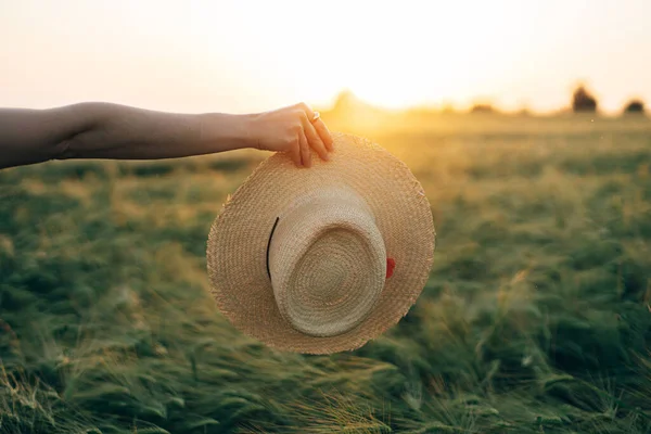 麦畑の背景に夕日の光の中で女性の手に帽子をストロー 夏の夜の田舎を楽しむスタイリッシュな女性 大気中の静かな瞬間 素朴な遅い生活 — ストック写真