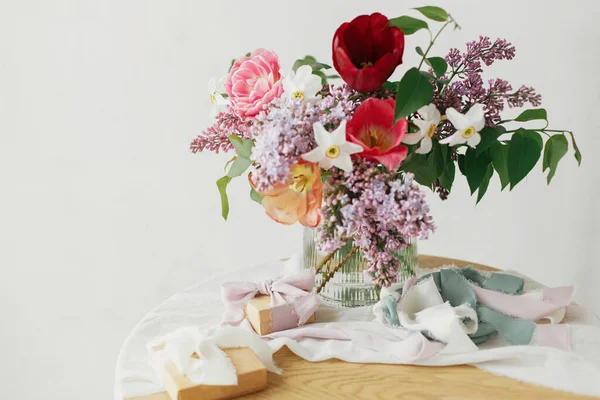 素朴な木製のテーブルの上にギフトボックスとスタイリッシュな花束 幸せな母の日 春のライラック チューリップ モダンな素朴な部屋で柔らかい生地に存在するダフォディルスの配置 幸せな女性の日 — ストック写真