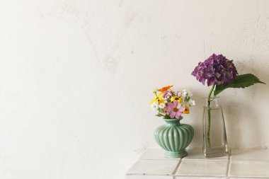 Mor ortanca ve kır arka planındaki fayans rafında kır çiçekleri. Bahçeden toplanan vazodaki güzel yaz çiçekleri, evdeki modern odada çiçek aranjmanları.