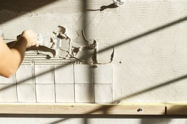 Modern kare fayanslar için duvara malayla yapıştırıcı döşeyen eller. İşçi güneş ışığında alçı duvara şık beyaz fayanslar yerleştiriyor. Yenileme