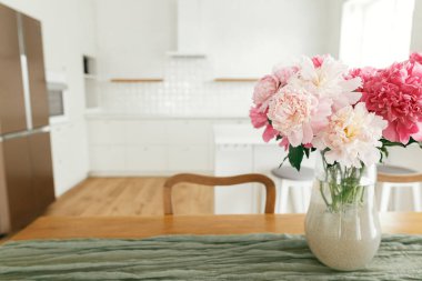 Ahşap masadaki vazodaki şakayıklar, adadaki şık beyaz mutfağın arka planı, ahşap raflar ve yeni evdeki aletler. Çiftlik evinde modern mutfak.