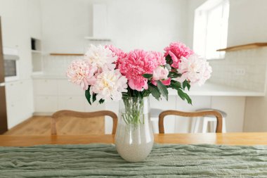 Yeni İskandinav evindeki aletlerle süslü beyaz mutfağın arka planında, ahşap masadaki güzel şakayıklar. Modern mutfak iç mimarisi ve yaz çiçekleri düzenlemesi