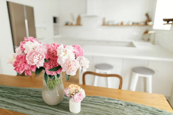 モダンなキッチンインテリア 新しい北欧の家で家電製品とスタイリッシュな白いキッチンを背景に木製のテーブルの上に花瓶の美しい牡丹 夏の花の配置 — ストック写真