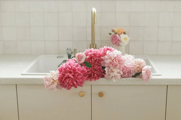 Beautiful peonies in sink on background of brass faucet and white counter in new scandinavian house. Pink peony and roses flowers in modern kitchen interior, summer floral arrangement