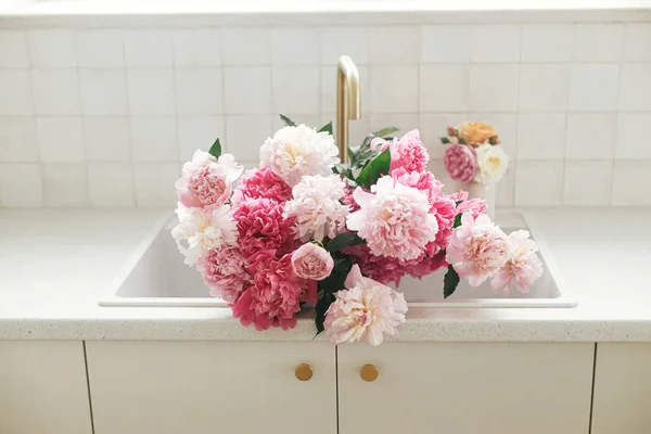 モダンなキッチンインテリア 農家で夏の花の配置でピンクの牡丹とバラの花 新しいスカンディナヴィアの家で真鍮の蛇口と白のカウンターを背景にシンクの美しい牡丹 — ストック写真