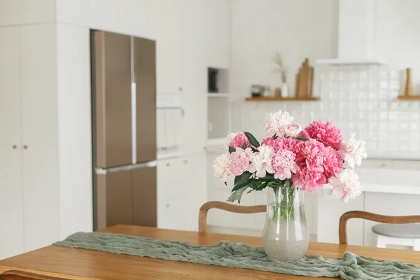 新しい北欧の家に家電製品とスタイリッシュな白いキッチンを背景に木製のテーブルの上に花瓶の美しい牡丹 モダンなキッチンインテリアと夏の花の配置 — ストック写真
