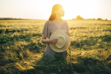 Çiçekli elbiseli güzel bir kadın gün batımında arpa tarlasında dikiliyor. Atmosferik sakin bir an, kırsal ve yavaş bir hayat. Hasır şapkalı şık bir kadın ve yaz akşamının tadını çıkarıyor.
