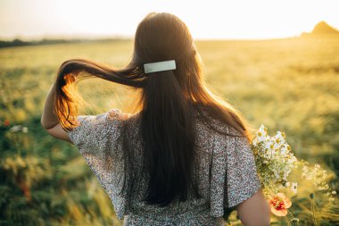 Arpa tarlasında günbatımının tadını çıkaran kır çiçekleriyle güzel bir kadın. Atmosferik sakin bir an, kırsal ve yavaş bir hayat. Tarz sahibi dişi kır çiçekleri toplayıp yaz akşamları dinleniyor.