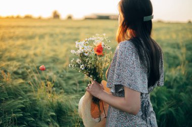 Gün batımında arpa tarlasında duran kır çiçekleriyle güzel bir kadın. Şık bir kadın yaz kırsalında dinleniyor ve çiçek topluyor. Atmosferik sükunet anı