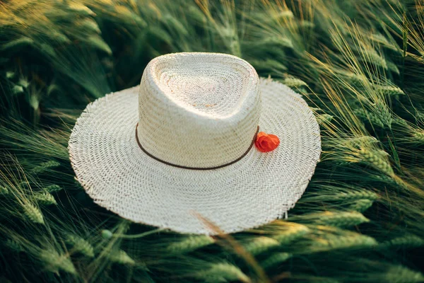 stock image Rustic straw hat and red poppy on barley ears in evening field close up. Wildflowers and farm hat in summer countryside. Atmospheric moment in evening meadow