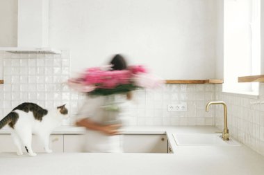 Yeni modern evdeki en az beyaz mutfağın arka planında kedisiyle şakayık düzenleyen bulanık bir kadın. Ev hanımlarının evi çiçeklerle dekore edişinin hareketli görüntüsü.