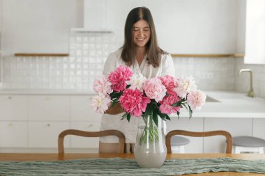 Yeni modern evin ahşap masasında vazoda güzel şakayıklar tutan bir kadın. En az beyaz mutfak arka planında çiçeklerle dekore edilen genç bayan yemek odası, taşınma ve ev temizliği