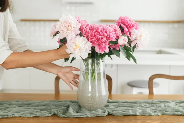新しい現代の家の木のテーブルの上に花瓶に美しい牡丹を配置女性 小さな白いキッチンを背景に花と若い女性の装飾ダイニングルーム に移動 — ストック写真
