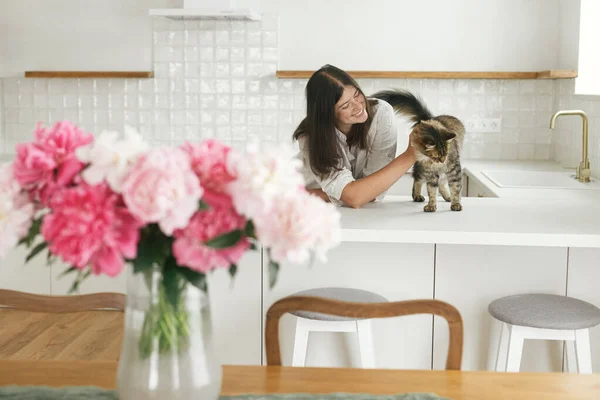 新しい現代の家で牡丹の花束と最小限の白いキッチンを背景にかわいい猫を愛撫幸せな女性 台所を掃除した後でリラックスしてペットと遊ぶ主婦 — ストック写真