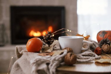 Cozy autumn. Warm cup of tea, pumpkins, autumn leaves, cones, cozy scarf on rustic wooden table on background of fireplace in farmhouse. Comfort fall in rural home. Happy Thanksgiving