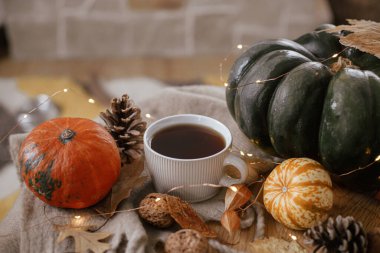 Warm cup of tea, pumpkins, autumn leaves, cones, cozy scarf and lights on rustic wooden table in farmhouse. Cozy fall in rural home. Happy Thanksgiving. Autumn hygge still life, banner