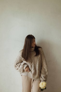 Sade örgü süveterli şık bir kadın kırsal otantik duvarın arka planında kasımpatı çiçeğiyle poz veriyor. Dengesiz düşüş. Hygge Sonbahar Evi 'nde genç ve şık bir bayan.