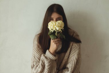 Sade örgü kazaklı şık bir kadın kırsal otantik duvarın arka planında kasımpatı ile poz veriyor. Dengesiz düşüş. Genç, modaya uygun, çiçekli yüzünü saklayan kadın portresi.