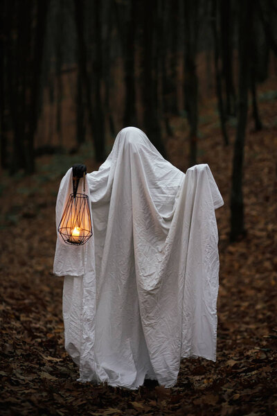 Spooky ghost holding glowing lantern in moody dark autumn forest. Person dressed in white sheet as ghost with light in evening fall woods. Happy Halloween! Trick or treat