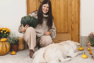 Güz balkabaklarıyla süslenmiş, kasımpatı ve çalılarla süslenmiş şirin bir köpekle oynayan örgü örülü süveter giymiş mutlu bir kadın. Evcil hayvan ve sonbahar düzenleme sahibi