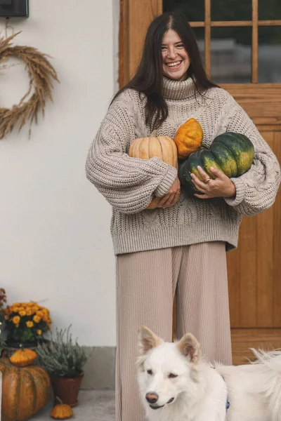 家の入り口の装飾のための大きい秋のカボチャを握る編まれたセーターの幸せな女性 彼女の犬の農場の玄関のドアで飾るスタイリッシュな女性 フォールアレンジメント — ストック写真