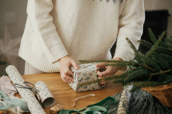 穿着时髦毛衣的女人把圣诞礼物用纸包在木制桌子上 在装饰过的丑闻室里装饰节日装饰品 圣诞快乐 手压得紧紧的 — 图库照片