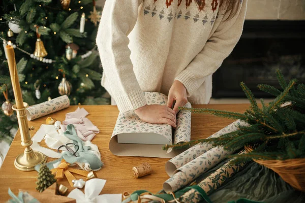 穿着时髦毛衣的女人把圣诞礼物用纸包在木制桌子上 在装饰过的丑闻室里装饰节日装饰品 圣诞快乐 手压得紧紧的 — 图库照片