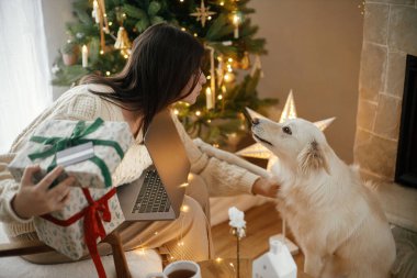 Mutlu kadın elinde kredi kartıyla şık Noel hediyeleri tutuyor ve Noel süslemeli Noel odasında ışıklarla sevimli beyaz köpekleri okşuyor. İnternetten Noel alışverişi ve Kara Cuma satışları.