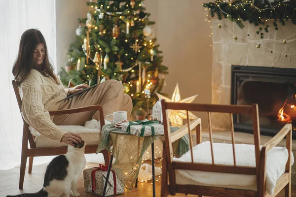 快乐的女人在笔记本电脑上工作 在装饰着圣诞树灯的圣诞节房间里爱抚可爱的小猫咪 圣诞节假期和宠物 寒假期间的远程工作 — 图库照片