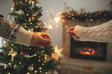 Modern ülke şöminesinin arka planında yanan maytaplar ve altın ışıklı noel ağacı. Mutlu yıllar! Havai fişekler ellerde parlıyor, çiftler şenlikli bir odada kutlama yapıyorlar.