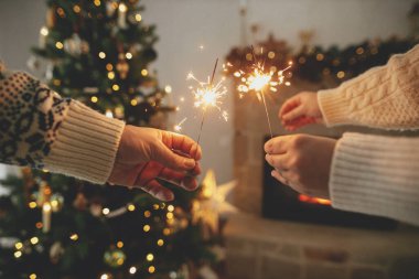 Modern şömine karşısında yanan havai fişekleri tutan eller ve altın ışıklı Noel ağacı. Mutlu yıllar! Arkadaşlar ve aile ellerinde yanan maytaplarla, atmosferik arifeyle kutlama yapıyorlar.