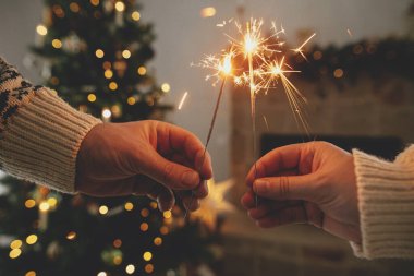 Modern şömine karşısında yanan havai fişekleri tutan eller ve altın ışıklı Noel ağacı. Mutlu yıllar! Arkadaşlar ve aile ellerinde yanan maytaplarla, atmosferik arifeyle kutlama yapıyorlar.