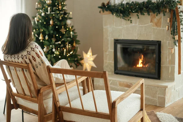穿着舒适毛衣的女人放松地坐在现代椅子上 看着身穿节日斗篷的壁炉 背景是装饰华丽的圣诞树 灯火通明 — 图库照片