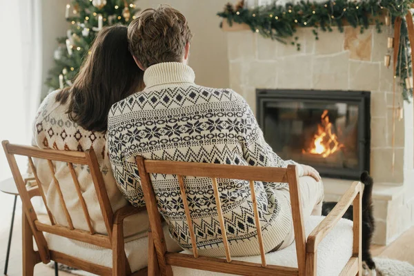 Stylish couple in cozy sweaters relaxing at fireplace with festive mantle on background of stylish decorated christmas tree with lights. Happy young family enjoying winter holidays