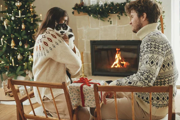 快乐的年轻家庭 穿着舒适的毛衣 抱着可爱的猫 在火堆前交换时尚的圣诞礼物 还有现代节日的斗篷和圣诞树 假期快乐 — 图库照片