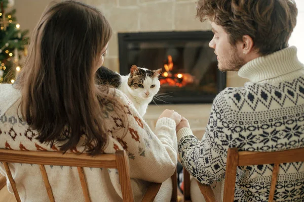Happy couple in cozy sweaters playing with cat and relaxing at fireplace with festive mantle and modern christmas tree with lights. Merry Christmas! Cozy winter