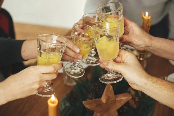 Hands with mimosa cocktail glasses clicking on background of stylish christmas table setting with fir branches, pine cones and candles. Friends and family celebrating and toasting with champagne
