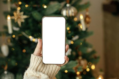 Noel telefonu maketi. Boş ekranlı akıllı telefonu altın ışıklı şık Noel ağacına karşı tutarak. Mesaj için yer var. Noel reklamı, akıllı telefon uygulama şablonu