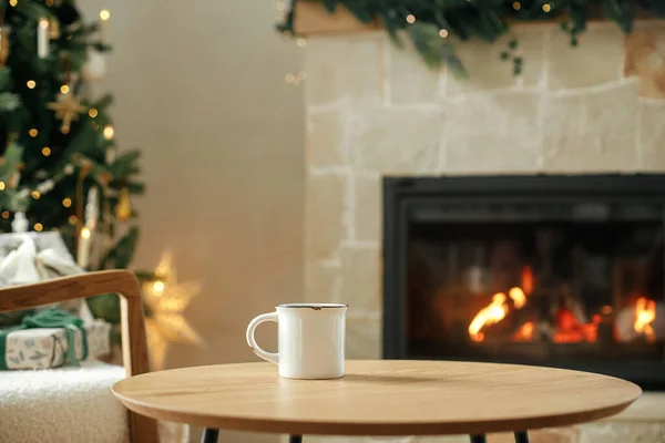 木制桌子上的老式白色杯子 与时尚的节日圣诞树 金色的灯光和壁炉相映成趣 杯模拟 圣诞节广告产品模板 案文的篇幅 — 图库照片