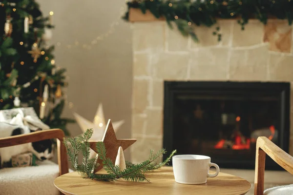 精美的白色杯子 桌上有木制星光装饰 与时尚的圣诞圣诞树相映成趣 还有金色的灯光和舒适的壁炉 杯模拟 圣诞节广告产品模板 案文的篇幅 — 图库照片