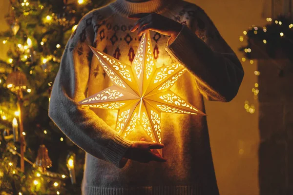夜の部屋で黄金のライトが付いているスタイリッシュな装飾されたクリスマス ツリーの背景に黄金の照らされたクリスマスの星を握る居心地の良いセーターの手 大気の魔法のイブ メリークリスマス — ストック写真