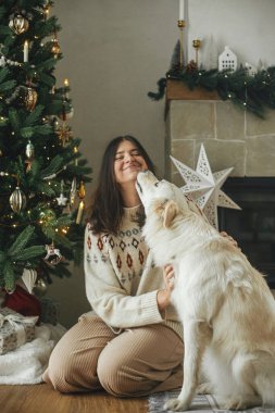 Şenlik odasındaki şık Noel ağacının arka planında sevimli beyaz köpekle sarılıp oynayan mutlu bir kadın. Kış tatili ve evcil. Mutlu Noeller.!