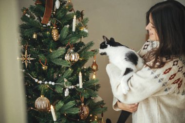 Şirin bir kedi tutan ve festival odasında şık bir Noel ağacı süsleyen rahat kazaklı mutlu bir kadın. Hayvan ve kış tatillerinin sahibi. Mutlu Noeller.!