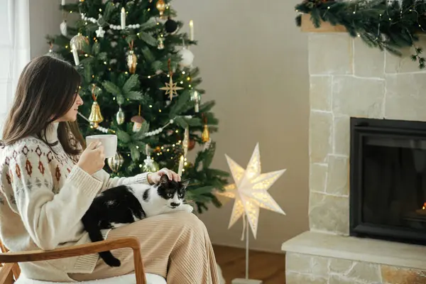 暖炉とスタイリッシュな装飾されたクリスマスツリーの背景にかわいい猫と暖かいお茶と座っている居心地の良いセーターの女性 ペットと衛生冬休みのオーナー メリークリスマス — ストック写真