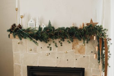 Noel dalları, çan çanları çanları, ahşap süsler ve ev dekorasyonuyla süslenmiş şömine rafı. Modern çiftlik evinin oturma odasındaki şenlikli kırsal Noel şöminesi