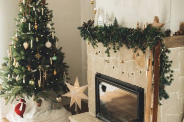 Şık dekore edilmiş şömine rafı, yılbaşı dalları, dekorasyonlar ve ışıklandırmalı modern Noel ağacının arka planında çanlar. Modern çiftlik evindeki kırsal Noel oturma odası