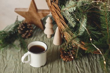 Şık bir fincan sıcak çay, sepetteki köknar dalları, tahta ağaçlar ve yıldız, kozalaklar ve masa ışıkları. Modern Noel rustik çevre dostu dekor ve kupa modeli, İskandinav kızlık zarı