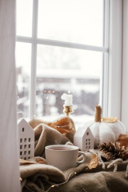 Cozy Autumn. Warm cup of tea, stylish pumpkin pillows, fall leaves, candle, lights and cute buildings decoration on brown scarf on windowsill. Autumn hygge, fall home decor