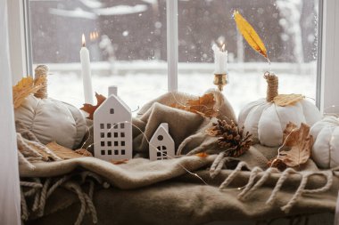 Cozy Autumn. Stylish pumpkin pillows, fall leaves, candle, lights and cute buildings decoration on brown scarf on windowsill. Autumn hygge, fall home decor. Happy Thanksgiving